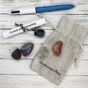May include: A collection of crystals and a pen arranged on a white wooden surface. A small beige drawstring bag with "Sentimental Gems" printed on it contains several polished stones. A scroll tied with twine reads "Crystals for Channeling."