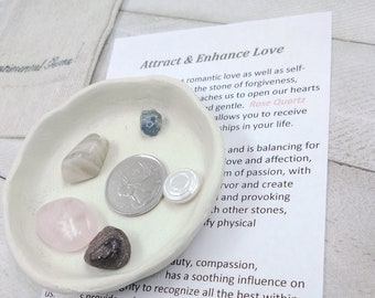 May include: A white ceramic dish with a variety of crystals and stones, including a pink rose quartz, a clear quartz, a blue sapphire, a grey agate, and a dark brown garnet. The dish is on a white surface with a card that reads "Attract & Enhance Love" and includes information about the crystals and their properties.