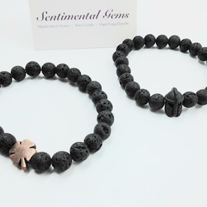 May include: Two black lava stone bead bracelets. One bracelet has a copper-coloured four-leaf clover charm, and the other has a black Spartan helmet charm. The bracelets are displayed on a white surface.