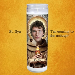 May include: A white glass prayer candle featuring an image of a man with curly hair, a brown robe, and a halo. The candle has the text "St. Ilya" and "I'm coming to the cottage."