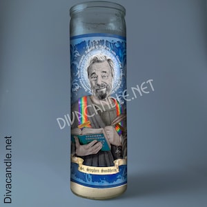 May include: A glass candle with a blue and white label featuring an illustration of Stephen Sondheim. The image includes text that reads "St. Stephen Sondheim" and "Sunday in the Park with George". The candle has a rainbow design.