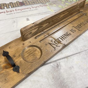 May include: A rectangular wooden serving board with a dark brown stained finish, featuring a black metal handle on the left side, a circular indentation, a raised back ledge, and the laser-engraved text "Nothing Is Impossible" on the surface.