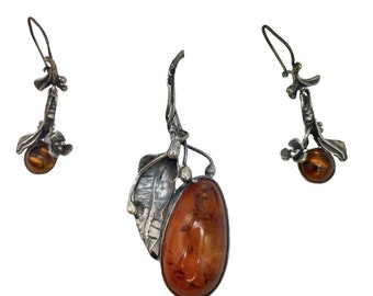 May include: A set of silver earrings and a pendant featuring amber stones. The earrings and pendant have a leaf design with a small amber stone at the bottom.
