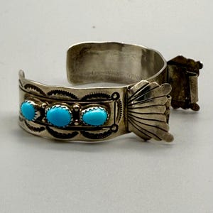 May include: A silver cuff bracelet with a hinged clasp and three turquoise stones. The bracelet is decorated with intricate silverwork.
