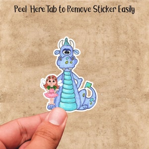 May include: A blue dragon with a green and blue striped body and a green tail stands next to a small fairy with pink wings and a pink dress. The fairy has a flower crown on her head. The sticker has the text "Peel Here Tab to Remove Sticker Easily" at the top.