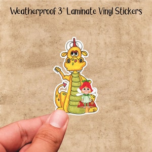 May include: A yellow dragon with a green body and red eyes is holding a small fairy with red hair and a green dress. The fairy is sitting on a red mushroom. The sticker is weatherproof and 3 inches in diameter.