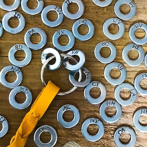 May include: A collection of silver metal washers, each stamped with a two-letter state abbreviation. Some washers are attached to a keyring with a yellow leather strap. The washers are scattered on a wooden surface.