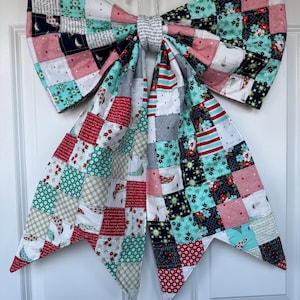 May include: A large, handmade fabric bow with a patchwork design. The bow features squares of various patterns, including sailboats, floral prints, and stripes, in shades of red, navy, teal, and pink. It is hanging on a white door.