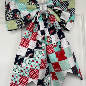 May include: A large, decorative bow made of patchwork fabric, featuring squares of red, white, navy, teal, and green patterns. The bow is hanging on a white door, with a white cord and black hook. The bow's center is wrapped in a patterned fabric.