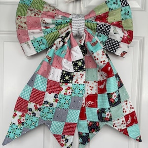 May include: A large, colorful quilted bow with a patchwork design. The bow features squares of red, pink, green, blue, and white fabric with floral and geometric patterns. The bow is hanging on a white door.
