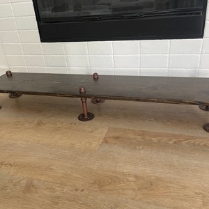 May include: A dark brown wooden shelf with copper pipe legs. The shelf is mounted to a white tiled wall.