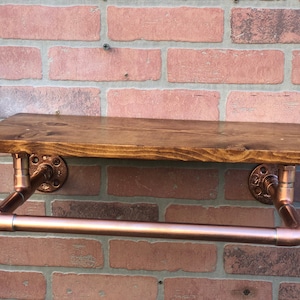 May include: A copper pipe shelf with a wooden shelf and a copper pipe towel bar. The shelf is mounted on a brick wall.