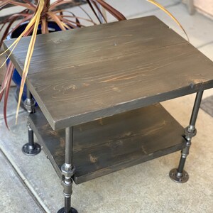 May include: A two-tiered wooden side table with black stained wood and black metal pipe legs.