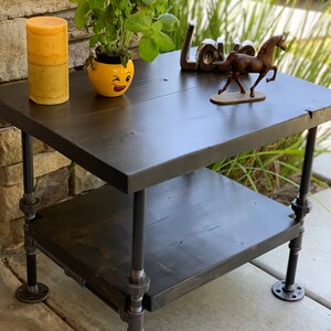 May include: A two-tiered black wooden side table with black metal pipe legs. The table has a yellow ceramic planter with a winking face on the top tier, and a small brown horse figurine on the bottom tier.