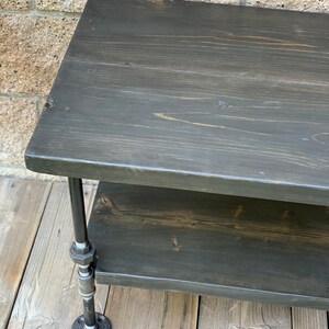 May include: A two-tiered wooden shelf with black stained wood and black metal pipe legs. The shelf is sitting on a wooden floor.