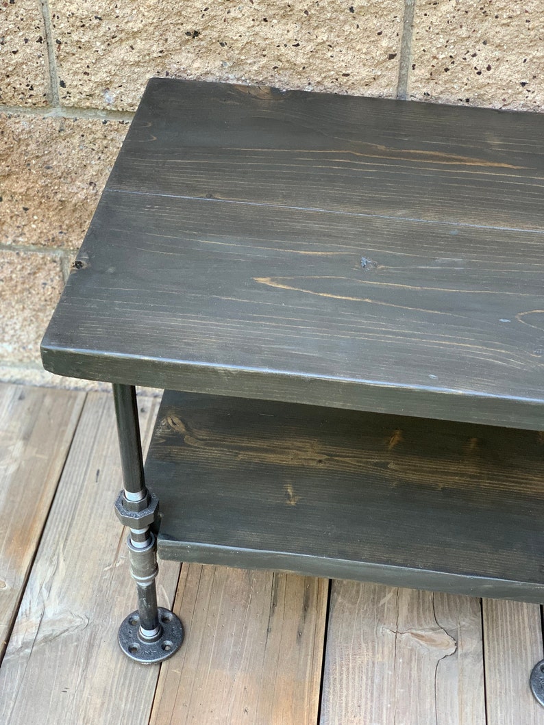 May include: A two-tiered wooden shelf with black stained wood and black metal pipe legs. The shelf is sitting on a wooden floor.