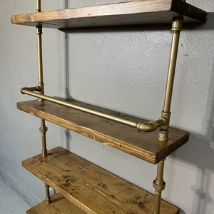 May include: A gold pipe shelving unit with four wooden shelves. The shelves are made of dark brown wood and have a rustic look. The unit is mounted to a wall and has a gold pipe rail for hanging items.