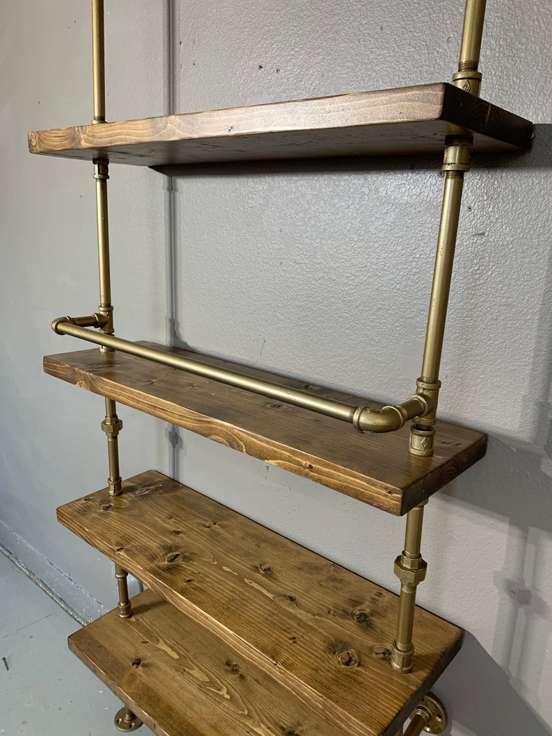 May include: A gold pipe shelving unit with four wooden shelves. The shelves are made of dark brown wood and have a rustic look. The unit is mounted to a wall and has a gold pipe rail for hanging items.