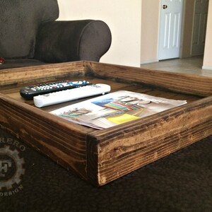 May include: A rustic wooden tray with a dark brown stain. The tray is sitting on a brown couch and has two remote controls and a magazine on top.