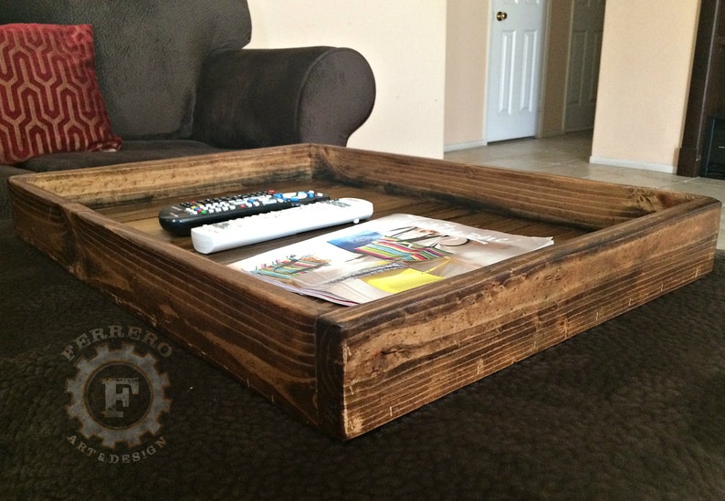 May include: A rustic wooden tray with a dark brown stain. The tray is sitting on a brown couch and has two remote controls and a magazine on top.