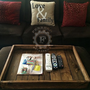 May include: A wooden tray with a dark brown stain. The tray has a magazine, two remote controls, and a decorative sign that says "Ferrero Art & Design" on it.