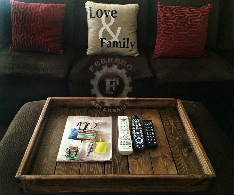 May include: A wooden tray with a dark brown stain. The tray has a magazine, two remote controls, and a decorative sign that says "Ferrero Art & Design" on it.