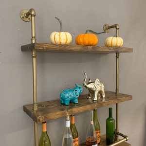 May include: A rustic wooden shelf unit with gold metal pipes and brackets. The shelves are decorated with pumpkins, decorative elephants, and wine bottles. The shelf unit is mounted on a wall.