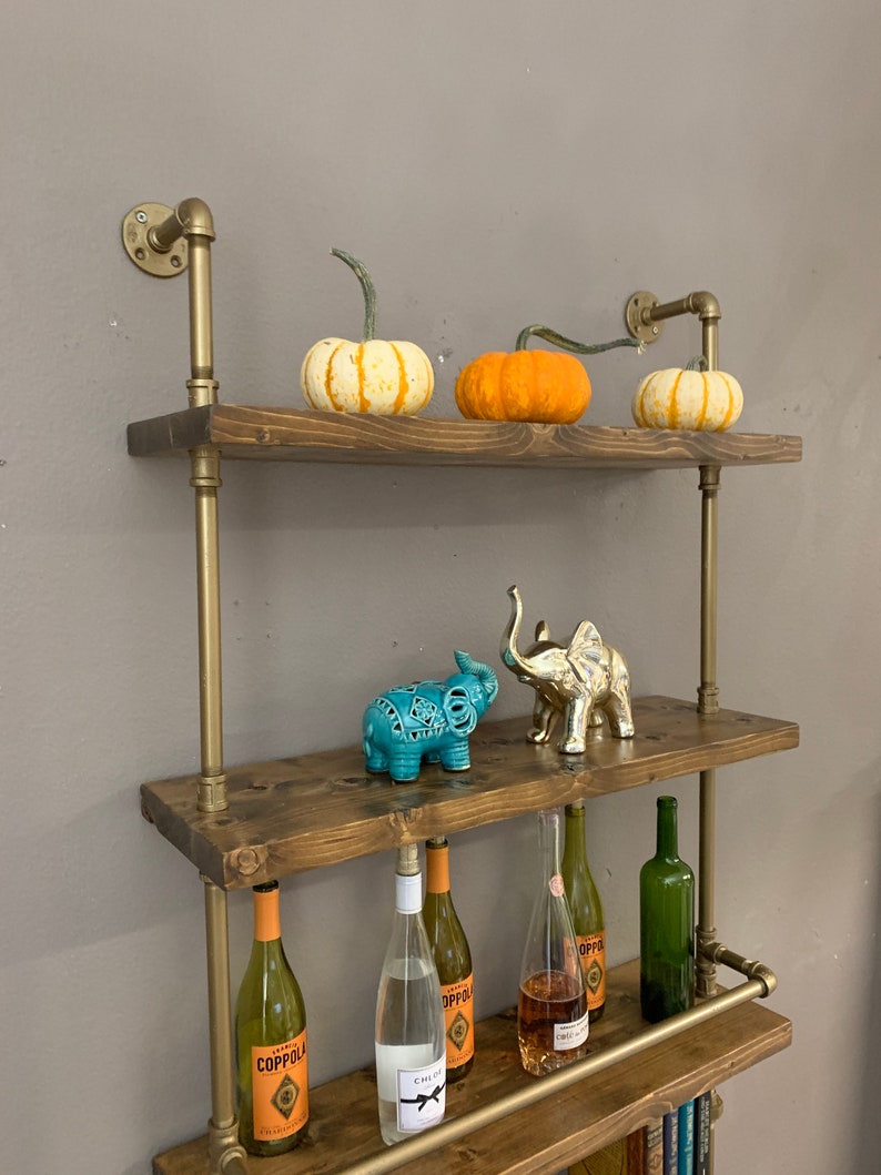 May include: A rustic wooden shelf unit with gold metal pipes and brackets. The shelves are decorated with pumpkins, decorative elephants, and wine bottles. The shelf unit is mounted on a wall.