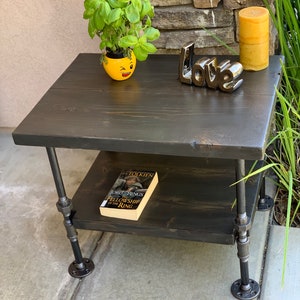 May include: A black wooden table with two shelves and black metal pipe legs. The table has a yellow ceramic planter with a green plant, a book titled "The Fellowship of the Ring" by J.R.R. Tolkien, and a black ceramic "Love" sign on top.