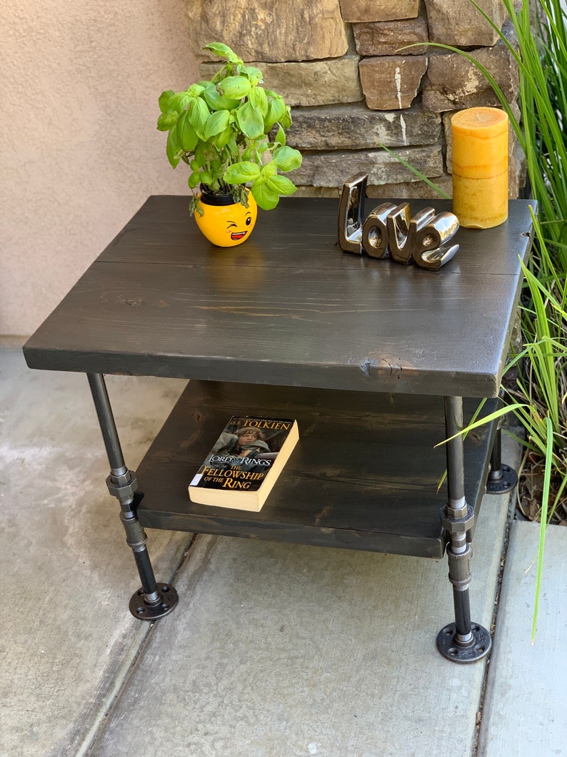 May include: A black wooden table with two shelves and black metal pipe legs. The table has a yellow ceramic planter with a green plant, a book titled "The Fellowship of the Ring" by J.R.R. Tolkien, and a black ceramic "Love" sign on top.