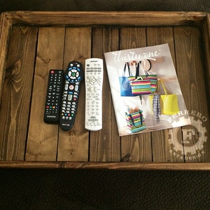 May include: A wooden tray with a dark brown stain. The tray has a raised edge and is made of several pieces of wood. There are three remote controls and a magazine on the tray. The magazine is titled "Thirty-one" and features colorful handbags. The tray has a white logo that says "Ferrero Art & Design".