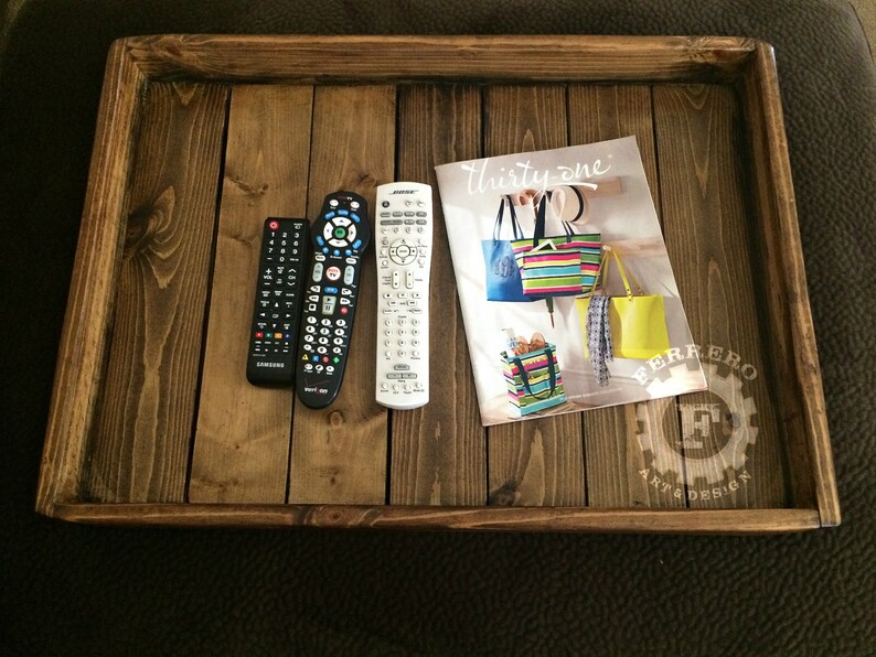 May include: A wooden tray with a dark brown stain. The tray has a raised edge and is made of several pieces of wood. There are three remote controls and a magazine on the tray. The magazine is titled "Thirty-one" and features colorful handbags. The tray has a white logo that says "Ferrero Art & Design".