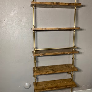 May include: A five-tiered wall shelf made of wood and gold metal pipe. The shelves are made of dark brown wood and the pipes are a shiny gold color. The shelf is mounted to the wall and has a rustic industrial style.