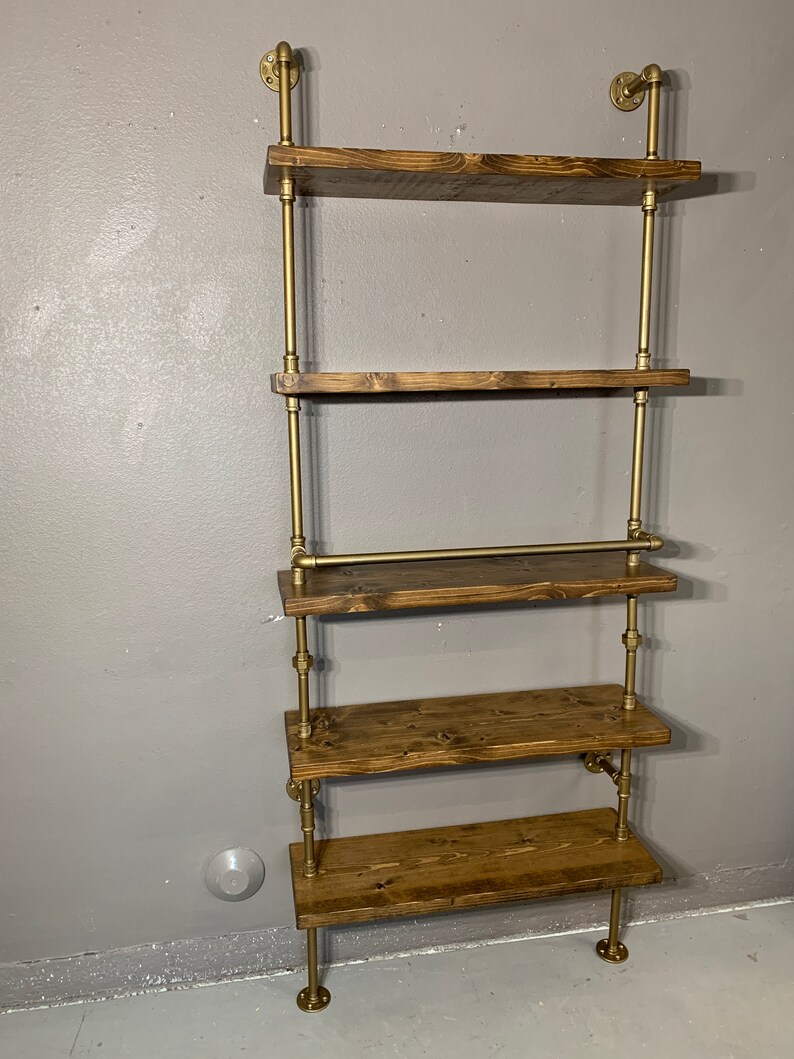 May include: A five-tiered wall shelf made of wood and gold metal pipe. The shelves are made of dark brown wood and the pipes are a shiny gold color. The shelf is mounted to the wall and has a rustic industrial style.