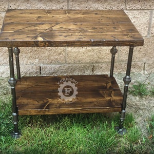 May include: A two-tiered wooden table with a dark brown stain. The table is made with industrial pipe legs and has a rustic, farmhouse style.