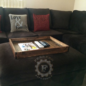 May include: A wooden tray with a rustic finish sits on a brown ottoman. The tray is filled with a magazine and two remote controls. The ottoman has a logo that reads "Ferrero Art & Design".