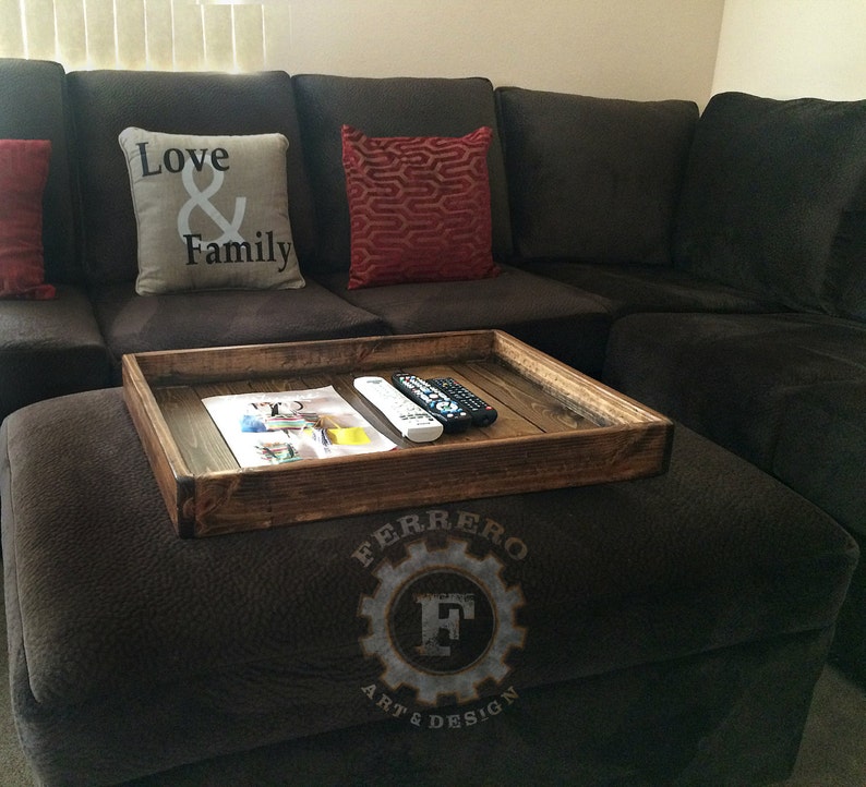 May include: A wooden tray with a rustic finish sits on a brown ottoman. The tray is filled with a magazine and two remote controls. The ottoman has a logo that reads "Ferrero Art & Design".