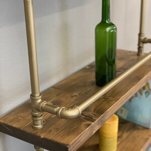May include: Gold pipe shelving unit with wooden shelves. The shelves are stained a dark brown and have a rustic look. A green glass bottle is sitting on the top shelf.