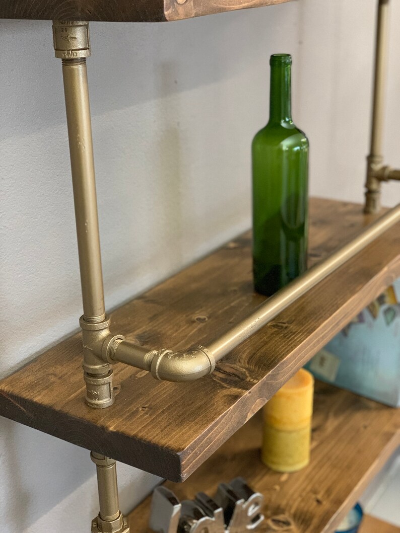 May include: Gold pipe shelving unit with wooden shelves. The shelves are stained a dark brown and have a rustic look. A green glass bottle is sitting on the top shelf.