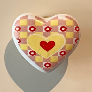 May include: A ceramic heart-shaped knob with a yellow and pink checkered pattern and a smaller red heart in the centre. The knob has a gold-coloured screw on the back.