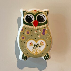 May include: A ceramic owl figurine with a green, yellow and white patterned design. The owl has large black eyes and a red beak.