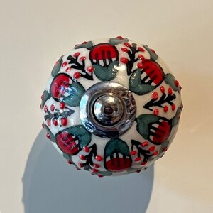 May include: White ceramic knob with a red and green floral design. The knob has a silver metal base.