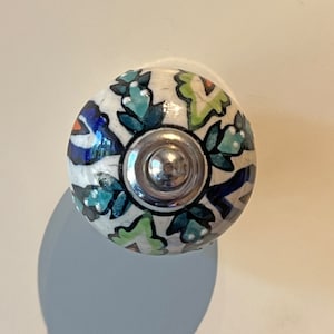 May include: A white ceramic knob with a blue, green and orange floral pattern. The knob has a silver metal centre.