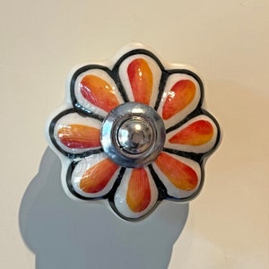 May include: A white ceramic flower-shaped knob with a silver centre and orange and yellow petals outlined in black.