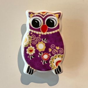 May include: A purple ceramic owl figurine with a floral pattern and black feet. The owl has large black eyes and a red beak.