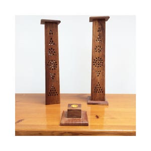 May include: Three wooden incense holders. Two tall, rectangular holders with decorative cut-out patterns stand on a wooden surface. A smaller square holder with a gold-coloured insert sits in front of them. The wood has a warm, brown tone.