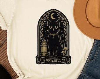 May include: Cream-colored t-shirt featuring a black cat design within an arched frame. The design includes a crescent moon, lantern, and the text "THE WATCHFUL CAT". A brown hat and blue jeans are visible.
