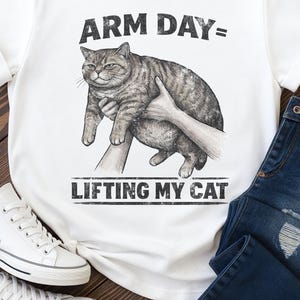 May include: White t-shirt with the text "ARM DAY= LIFTING MY CAT" in black. The graphic shows a tabby cat being lifted by two hands. The shirt is paired with blue jeans and white sneakers.