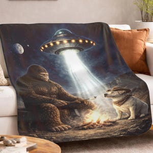 Peut inclure: Une couverture en tissu avec une scène fantaisiste : un Bigfoot grillant des guimauves avec un chien portant un chapeau de cowboy, sous un OVNI. Le fond est sombre, avec une lune et des étoiles.