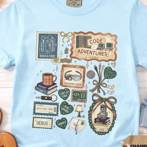 May include: Light blue t-shirt with a graphic design featuring computer-related imagery and text. The design includes the words "CODE ADVENTURES," "DEBUG," and "DEBUGGING VISION." The shirt is made of a soft material and has a crew neck.